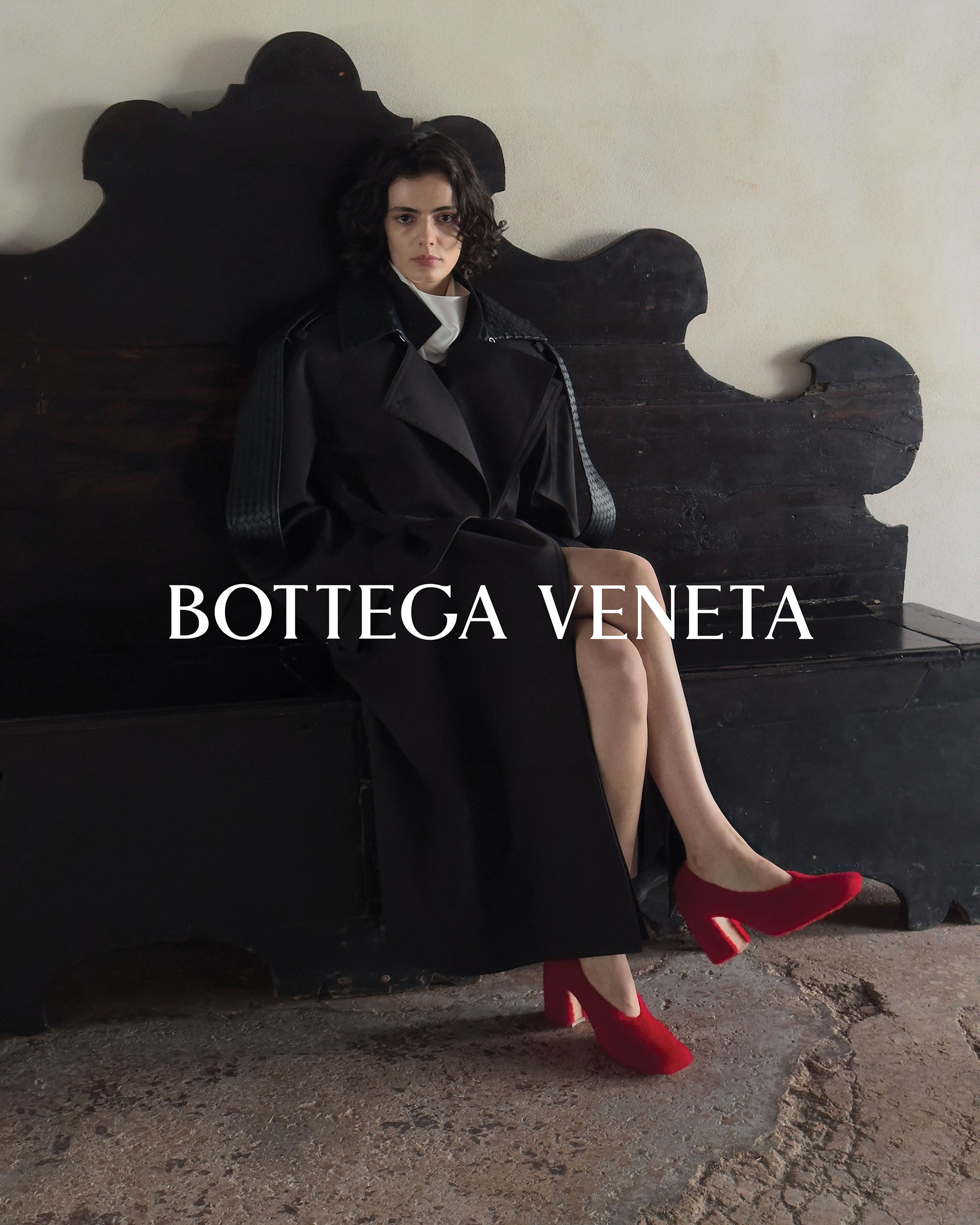Model seated on an ornate dark bench wearing a long black coat and red heels for Bottega Veneta