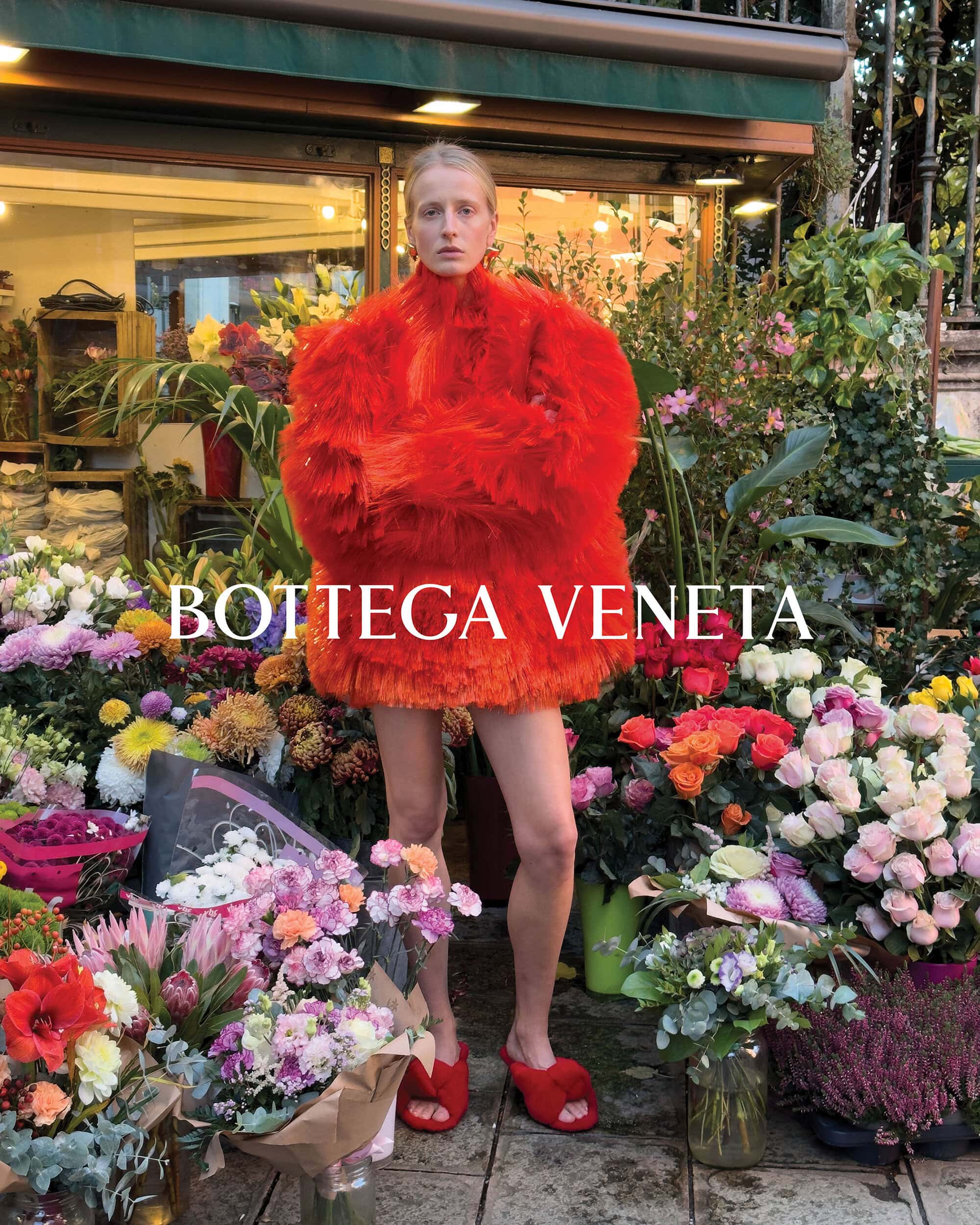 Fashion shot of a blonde woman wearing a fuzzy red dress standing in front of a flower stall with assorted colorful flowers.