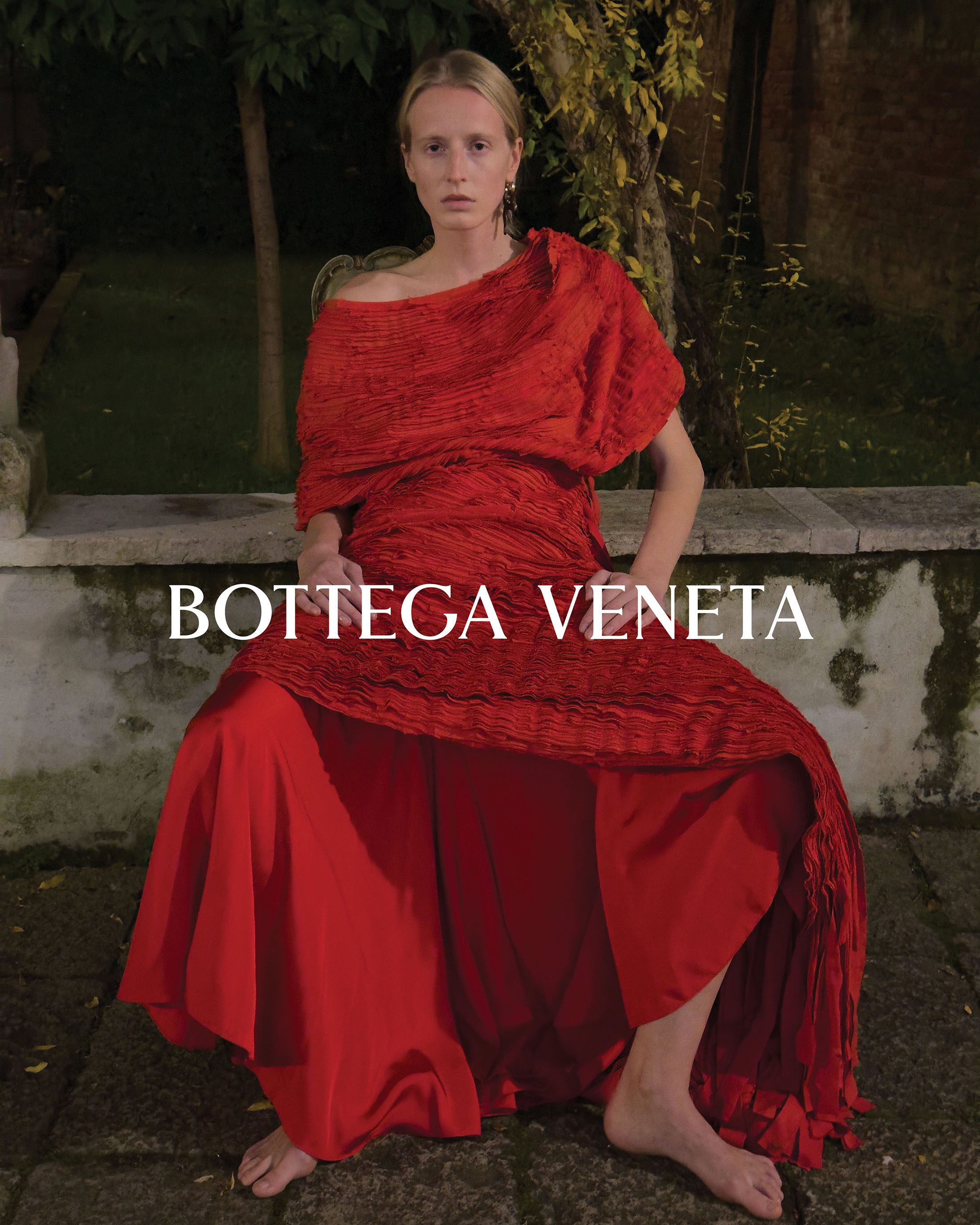 A fair-skinned female model in a dramatic red dress poses in an outdoor setting for a Bottega Veneta fashion campaign with visible logo.