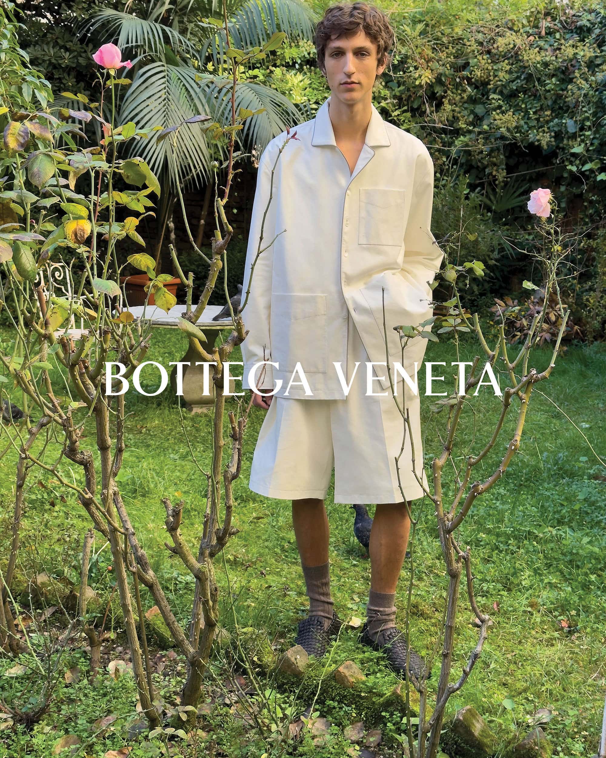 A young man in a white suit stands in a verdant garden, showcasing a Bottega Veneta fashion campaign with natural light and gentle floral backdrop.