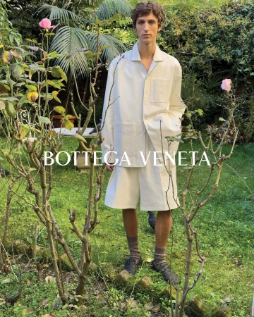 A young man in a white suit stands in a verdant garden, showcasing a Bottega Veneta fashion campaign with natural light and gentle floral backdrop.