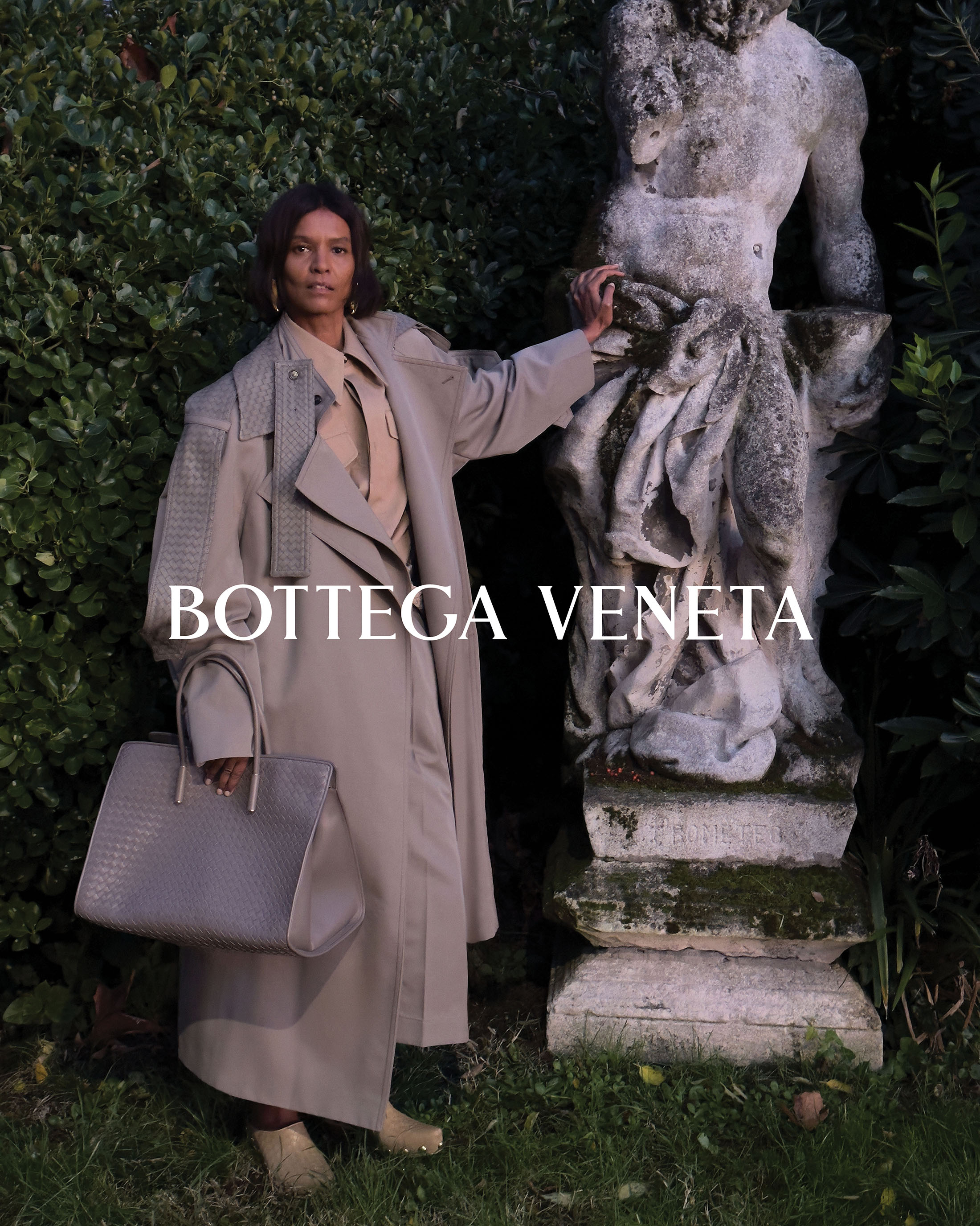 A woman poses in a trench coat with a handbag beside a weathered sculpture outdoors. Fashion photography showcases neutral tones and natural light.