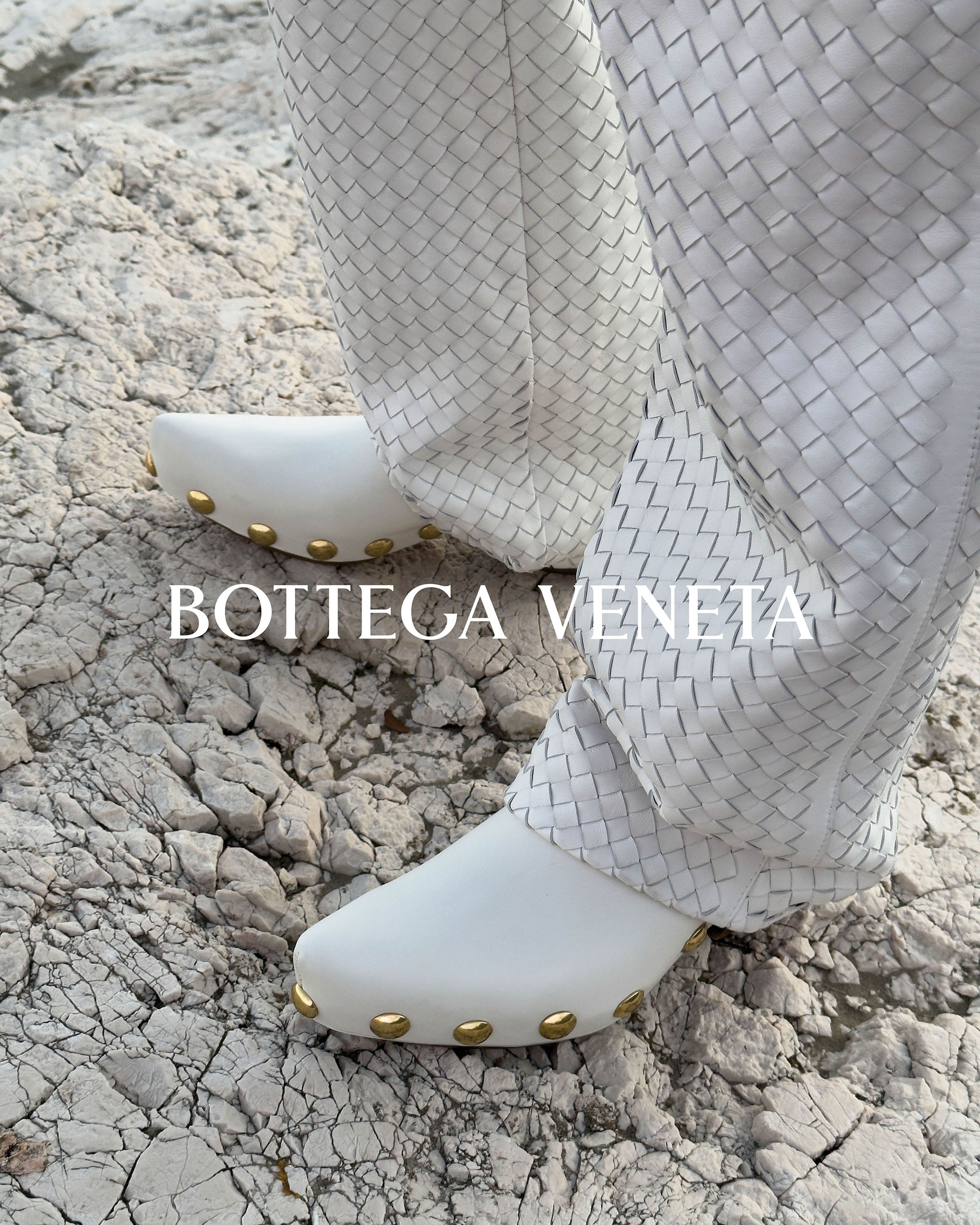 Close-up of white heeled clogs with gold accents and Bottega Veneta woven-texture pants standing on cracked white rocks. Fashion aesthetic.