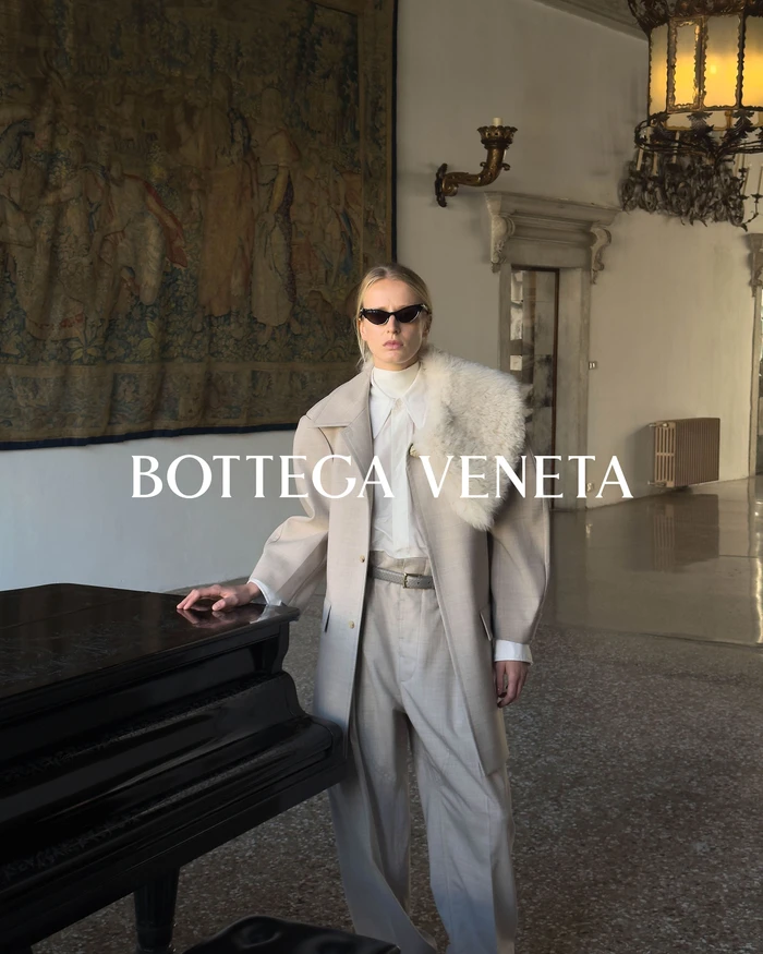 Model in cream coat and sunglasses standing next to a black piano in a grand hall with a large tapestry