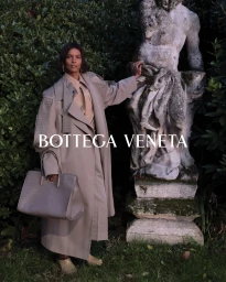 Model standing outdoors next to a classical stone statue wearing a beige trench coat and holding a structured grey handbag