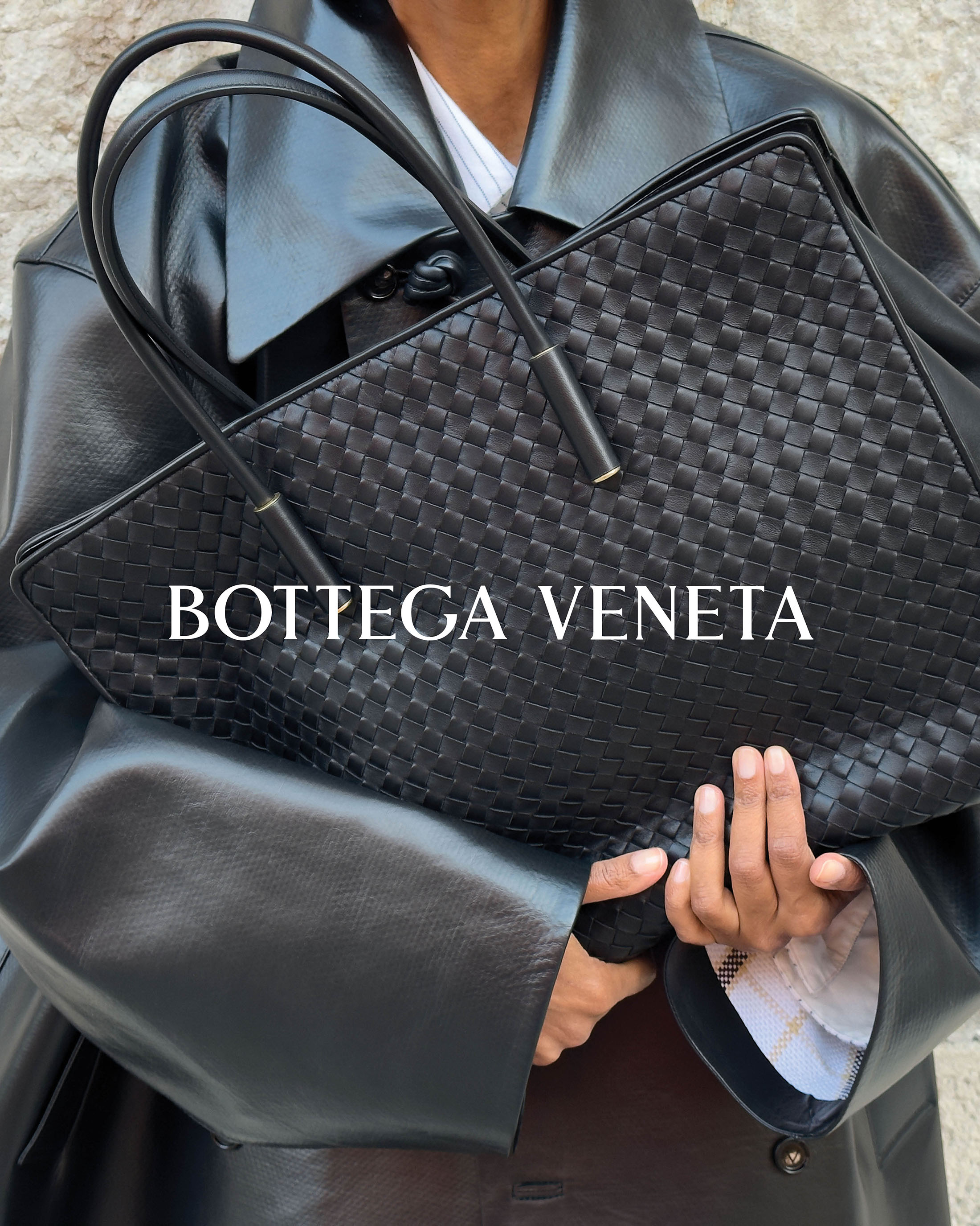 Model holding a black woven leather tote bag wearing a black leather coat for Bottega Veneta