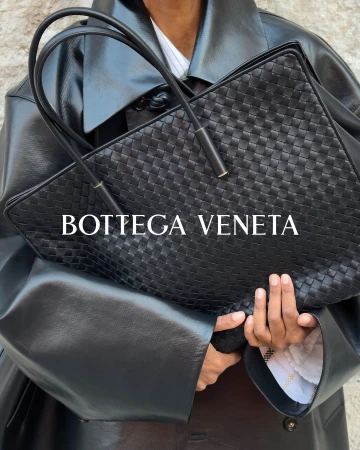 Model holding a black woven leather tote bag wearing a black leather coat for Bottega Veneta