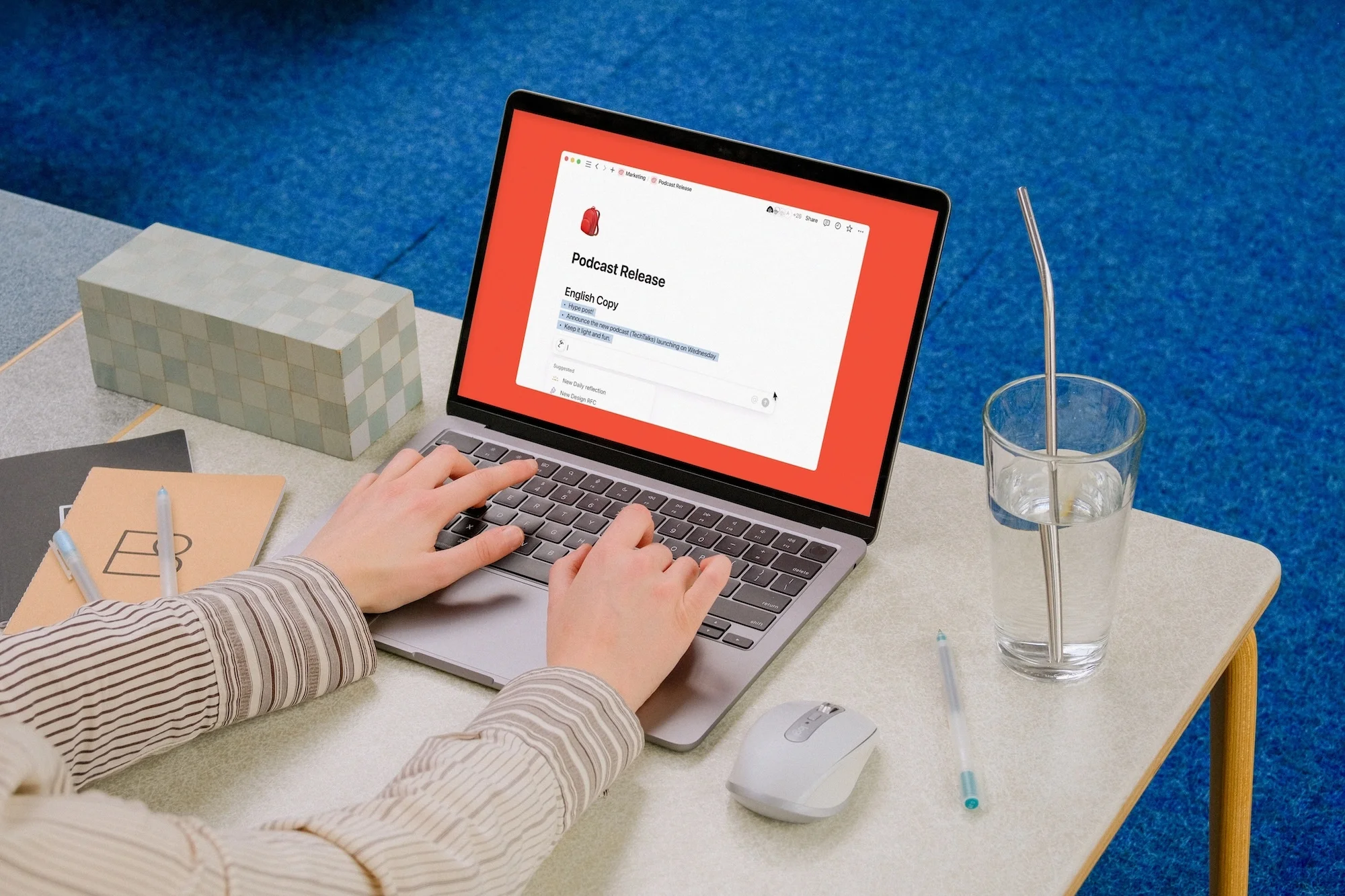 Person typing on a laptop with a podcast release document on screen, a glass of water, and a notebook on a desk with blue carpet.