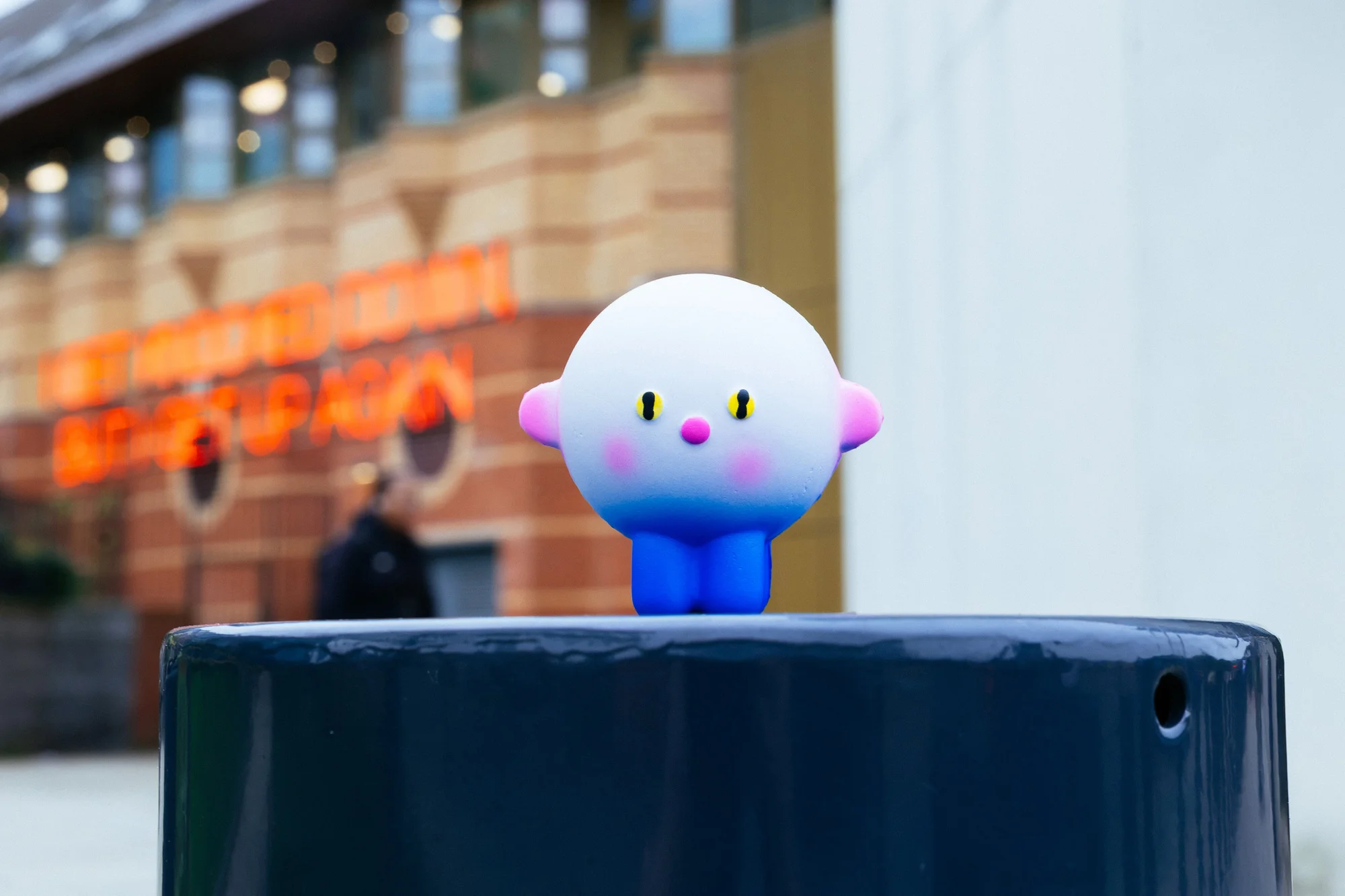 Whimsical toy figure with gradient white-to-blue body, pink cheeks, rests on a dark grey post outdoors, with a building in the background.