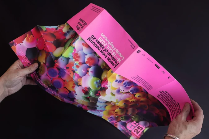 Hands holding a folded poster advertising Leeds International Festival of Ideas 2025 at Leeds Playhouse; pink, colorful, and modern.