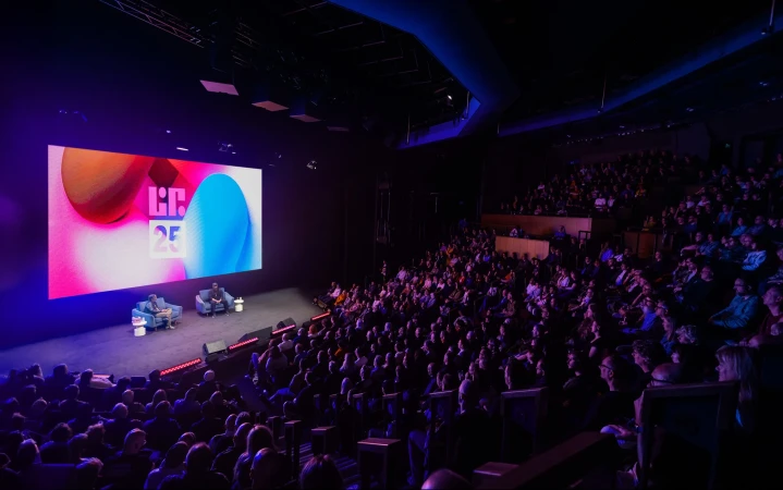 Large audience watches two men in chairs on stage in auditorium, illuminated by pink and blue lights with text on screen.