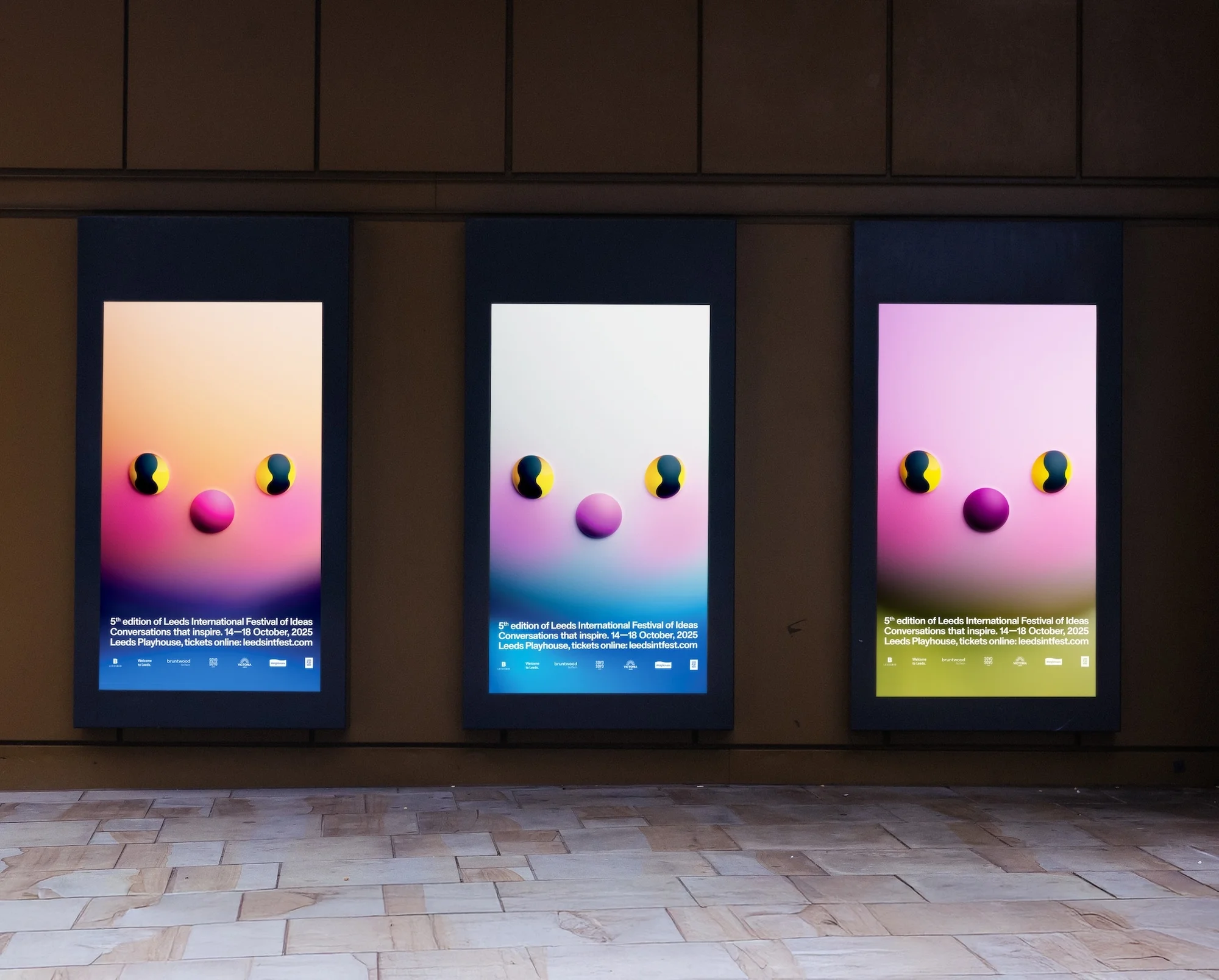 Three digital billboards display graphic art with pink and black circles on pastel backgrounds in a neutral-toned indoor space.