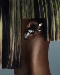 Close-up portrait of a person wearing Nothing Ear earbuds and metallic hoop ear cuffs under a blunt fringe haircut against a neutral background.