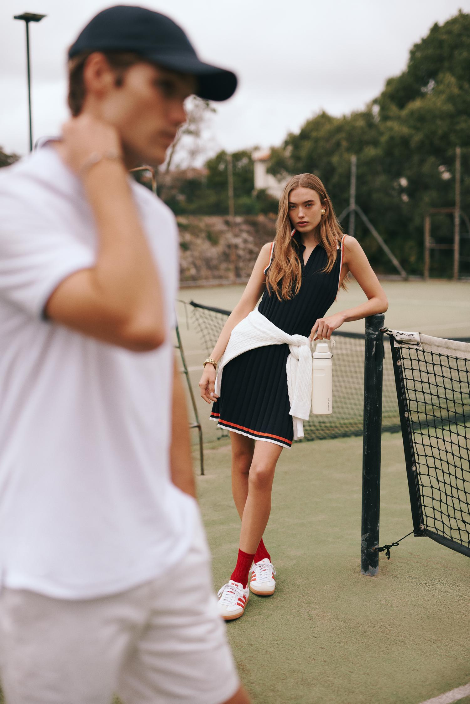 A woman in a black tennis dress poses on a tennis court with a man in a white polo shirt and baseball cap in the foreground.
