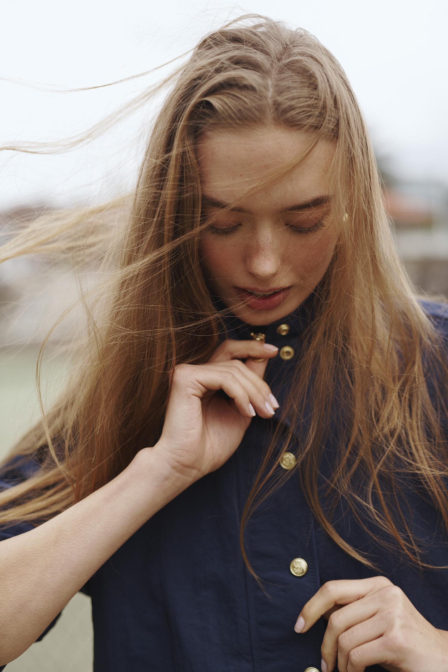 Portrait of an adult woman with long blonde hair, wearing a collared dark blue shirt with gold buttons, looking down outside.