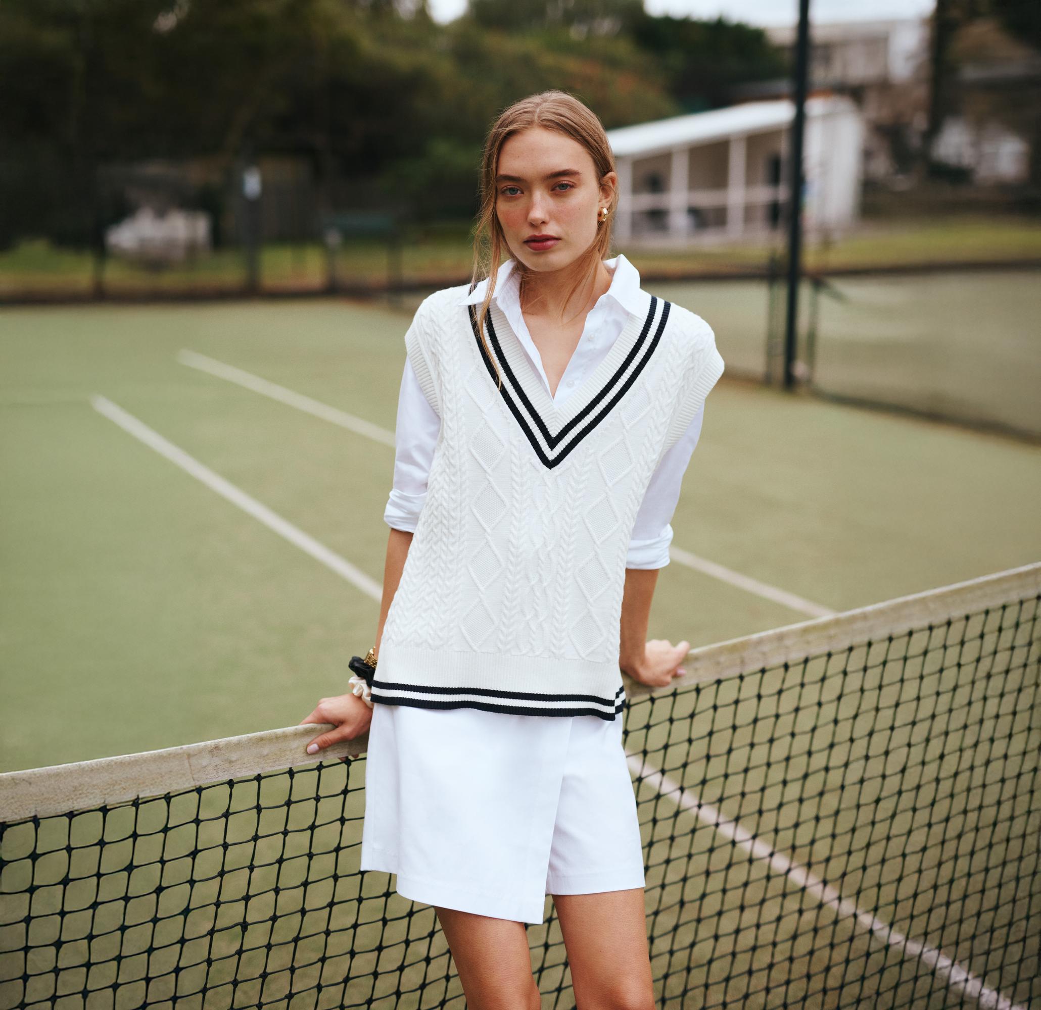 Young woman poses in preppy white shorts and sweater vest leaning on a tennis net on a green tennis court in an overcast outdoor setting.
