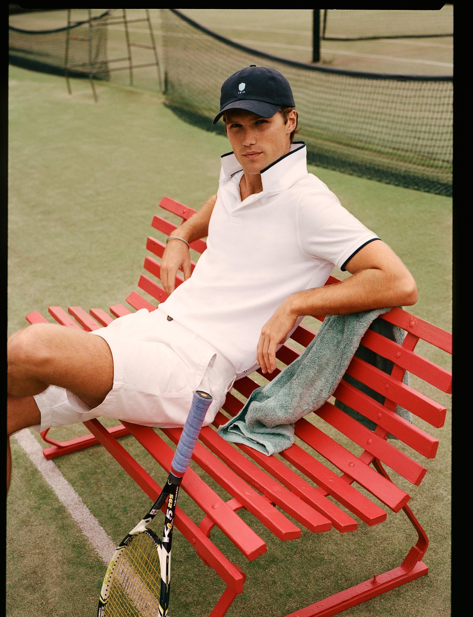 Male model poses on a red bench at a tennis court. He wears a white polo shirt, shorts, and a blue baseball cap. Athletic fashion.