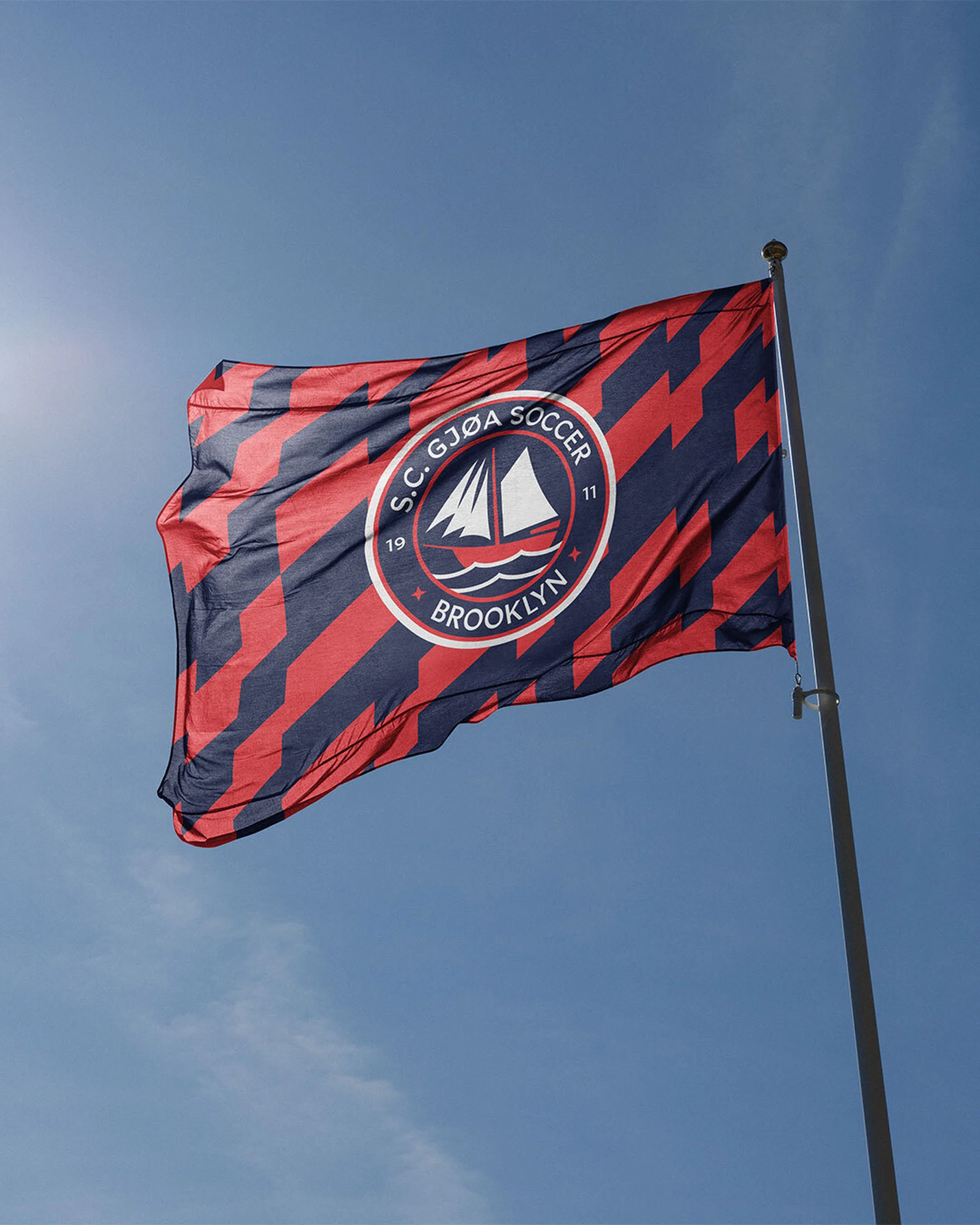 S.C. Gjoa Soccer flag featuring a sailboat logo against a clear blue sky on a sunny day with a modern graphic pattern.