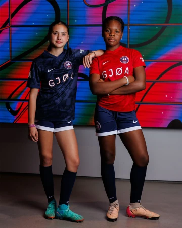 Two female soccer players in team uniforms stand shoulder-to-shoulder against a colorful geometric background in an indoor setting.