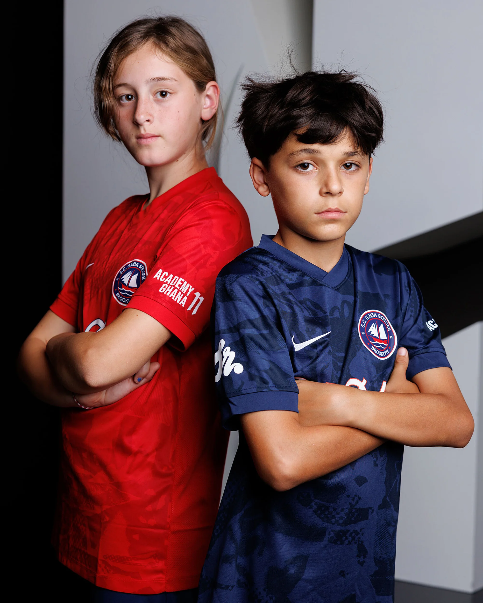 Two young soccer players in team jerseys stand back to back, arms crossed, indoors. The boy on the left wears a red jersey, the other a blue one.