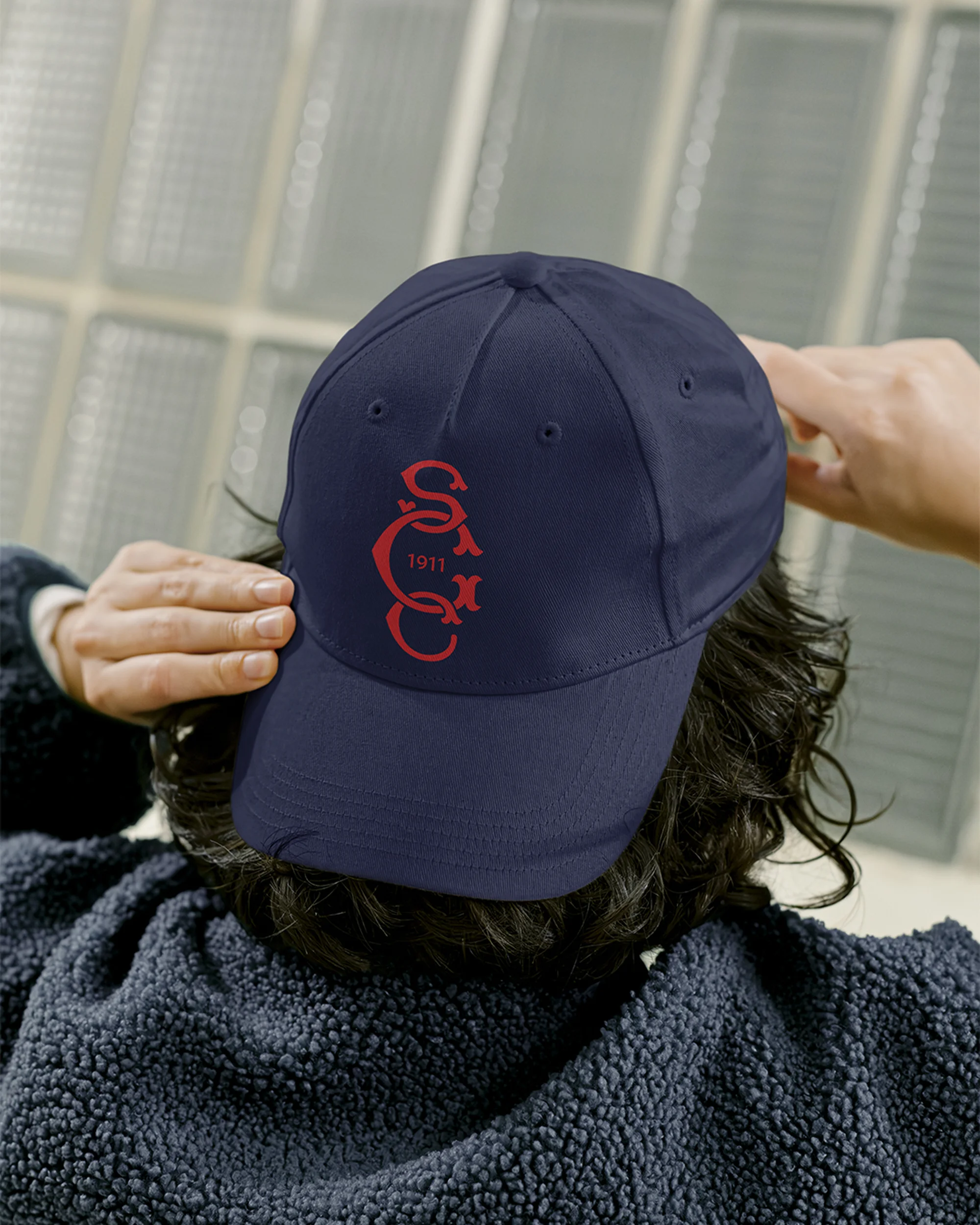 Close-up of a dark blue baseball cap with a red logo being worn by a person in a navy fleece jacket against a neutral background.
