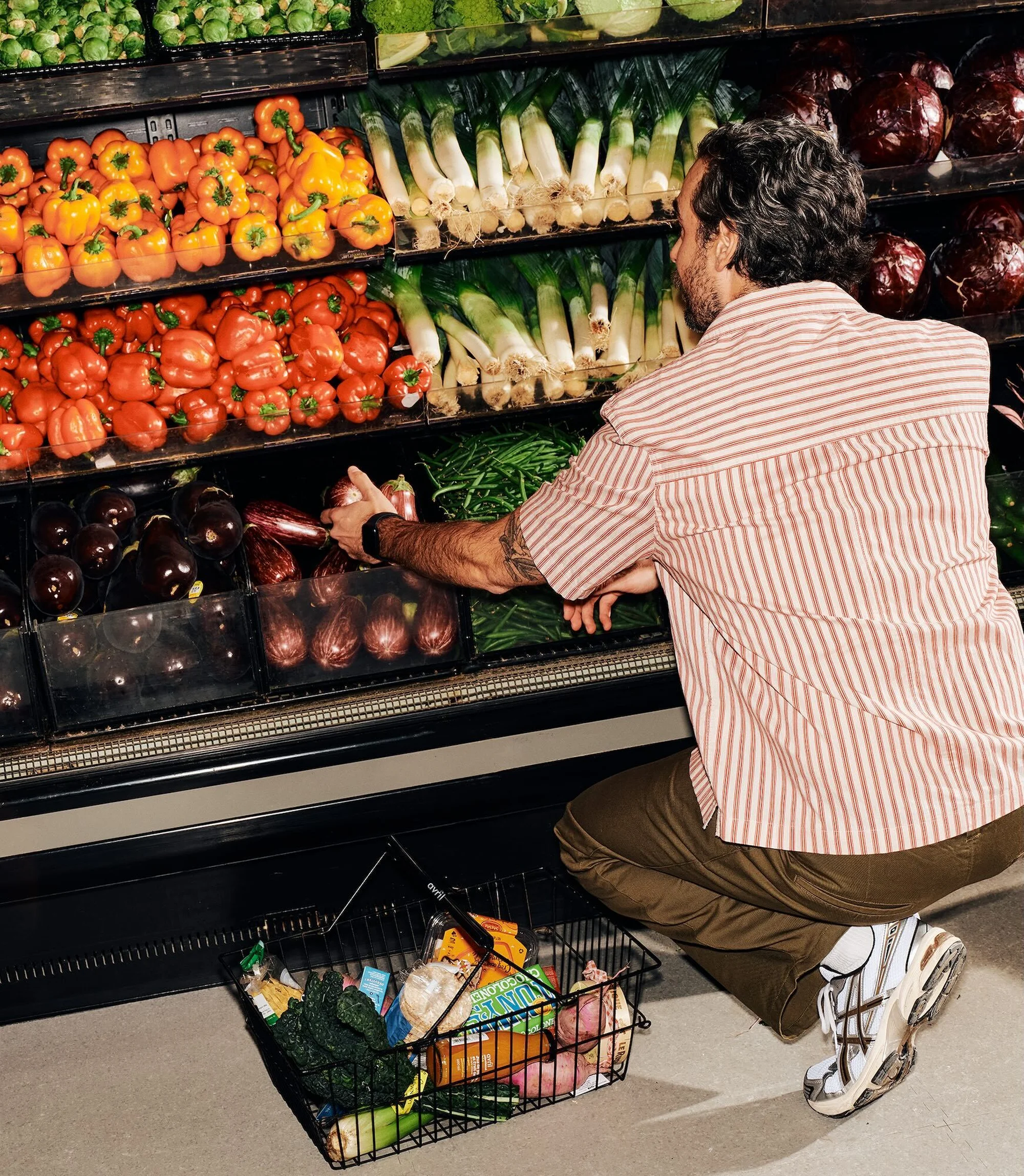 An adult male squats to select eggplants in a grocery store produce section, holding a black wire shopping basket full of fresh produce.
