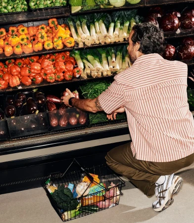 An adult male squats to select eggplants in a grocery store produce section, holding a black wire shopping basket full of fresh produce.
