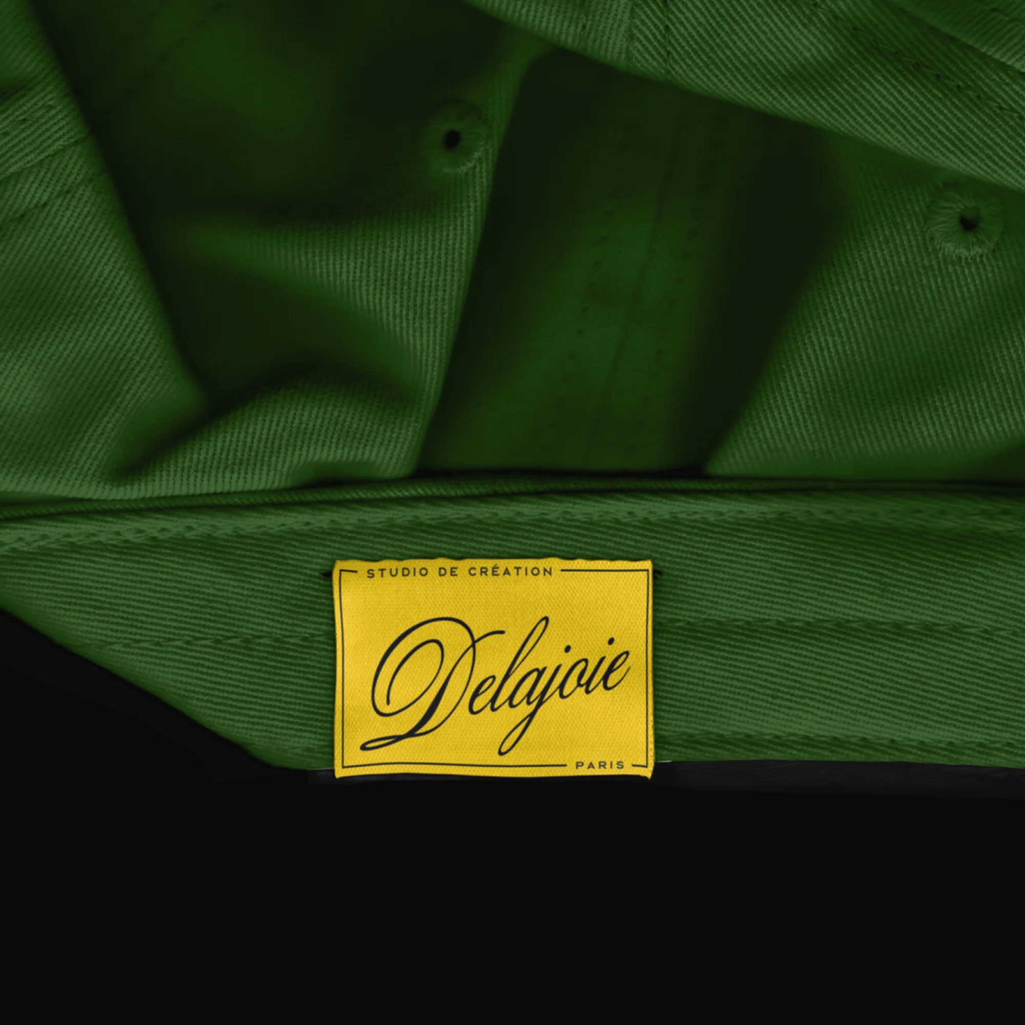 Close-up of a green baseball cap with a yellow tag featuring black script that reads 'Studio de Creation Delajoie Paris'.