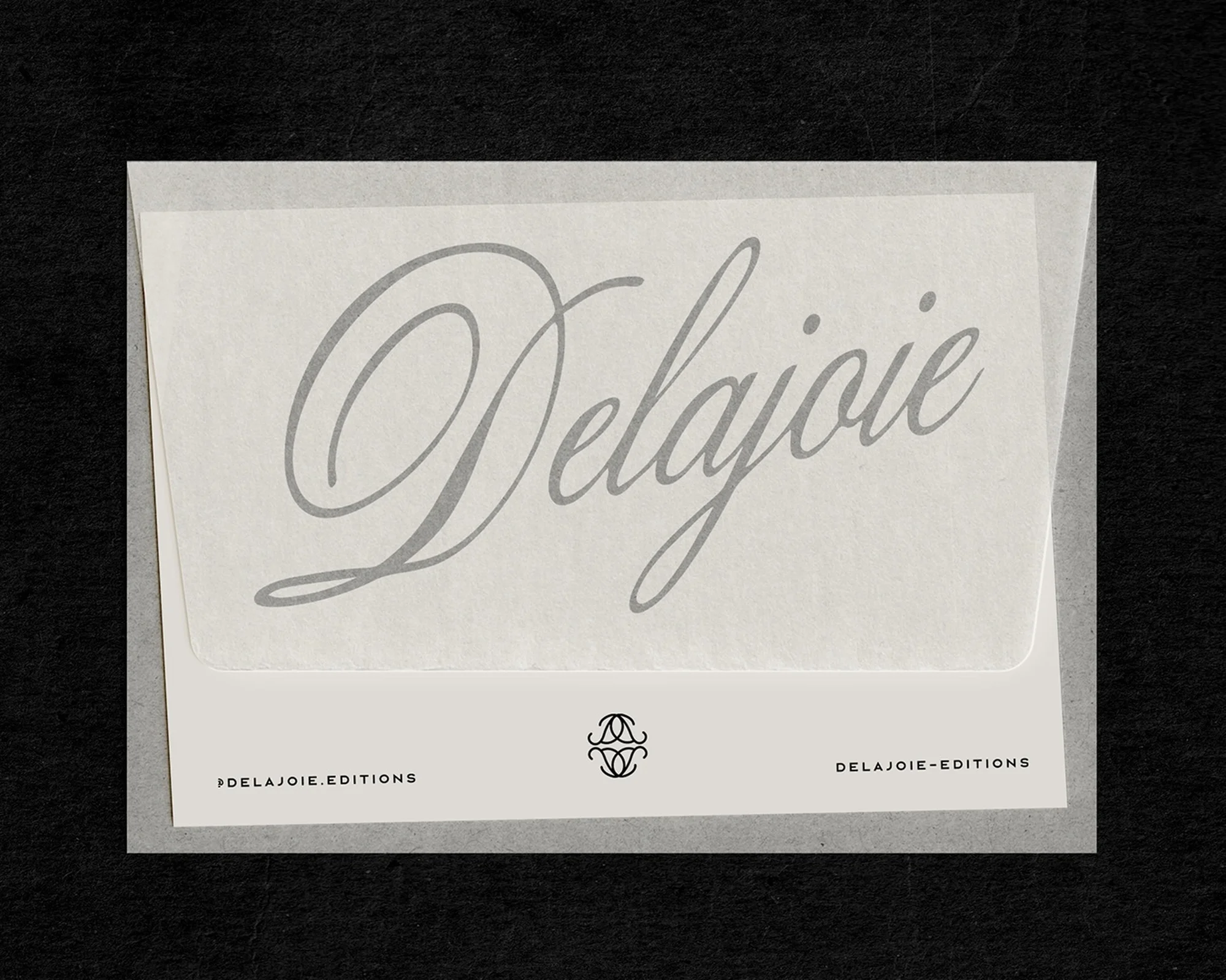 Close-up studio shot of white paper envelope with 'Delajoie' typography in muted gray, against a black background. Minimalist aesthetic.