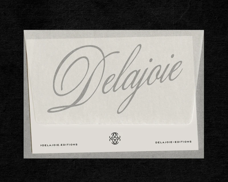 Close-up studio shot of white paper envelope with 'Delajoie' typography in muted gray, against a black background. Minimalist aesthetic.