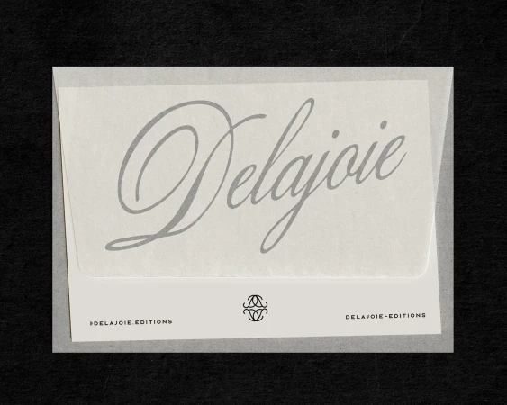 Close-up studio shot of white paper envelope with 'Delajoie' typography in muted gray, against a black background. Minimalist aesthetic.