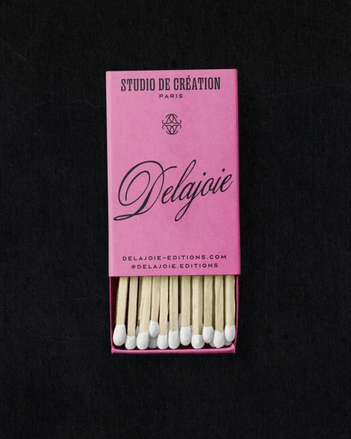 Close-up of a pink matchbox with 'Delajoie' written on it and wooden matches with white tips, placed on a dark, textured surface.