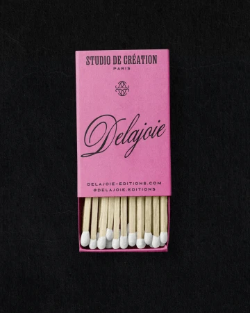 Close-up of a pink matchbox with 'Delajoie' written on it and wooden matches with white tips, placed on a dark, textured surface.