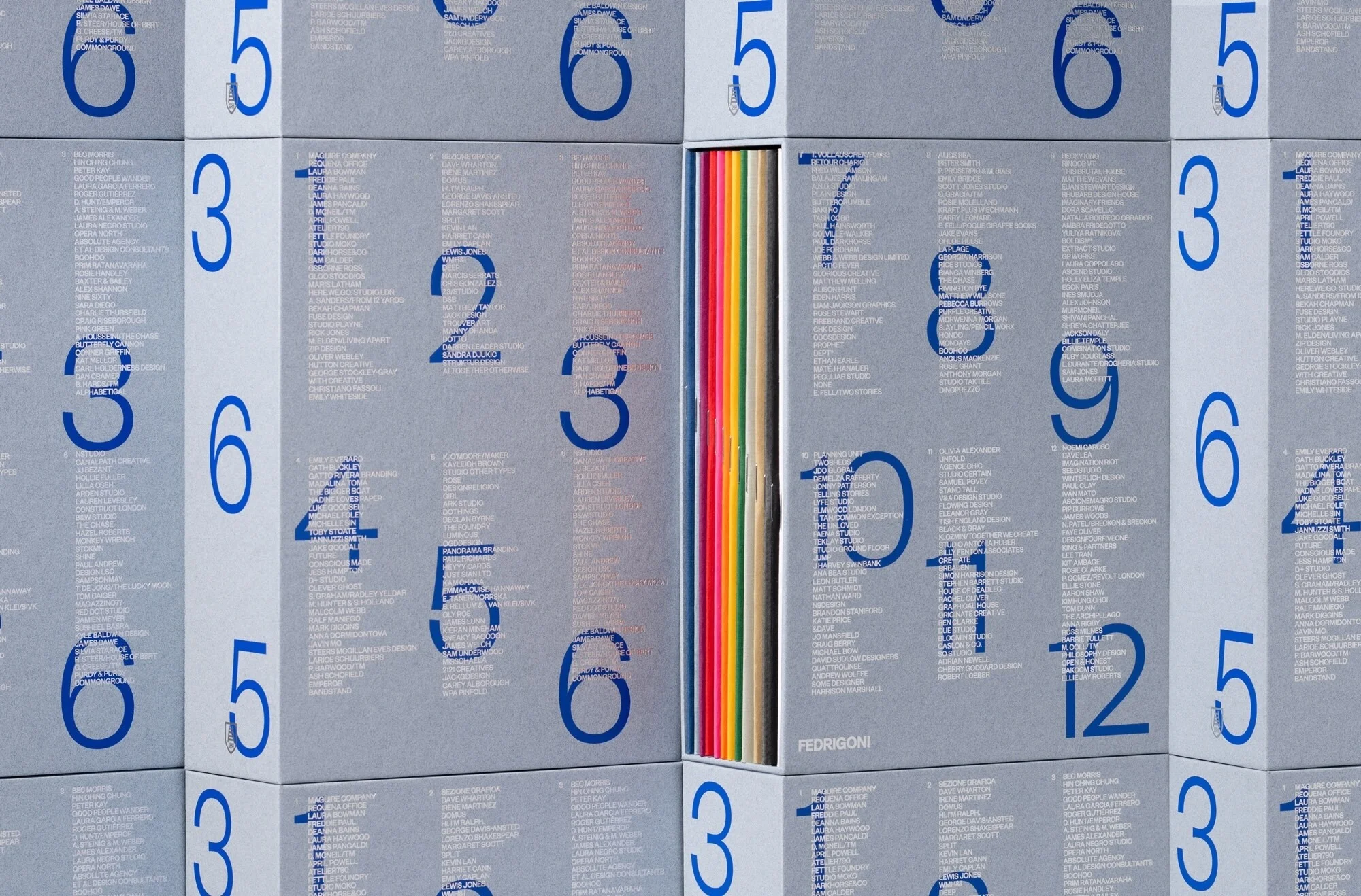 Close-up of stacked Fedrigoni paper sample books with blue numbers and text on gray covers and a single open book with colorful papers.