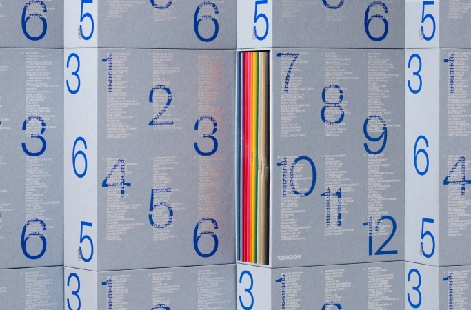 Close-up of stacked Fedrigoni paper sample books with blue numbers and text on gray covers and a single open book with colorful papers.