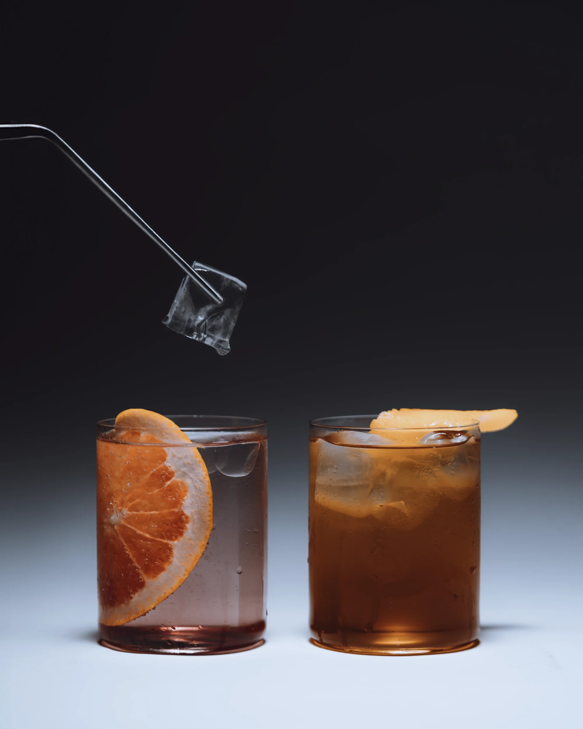 Two cocktails, one pink and one amber, are garnished with citrus. Metal tongs hold an ice cube over the pink drink on a gray surface.