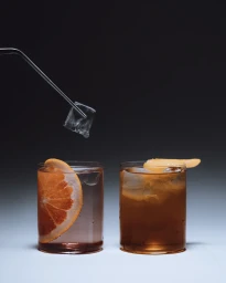 Two cocktail glasses with citrus slices against a dark gradient background as ice is dropped by metal tongs
