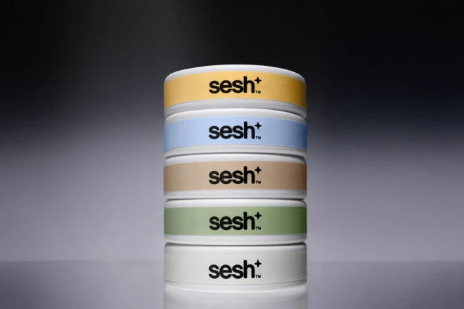Stack of five white containers with pastel color labels and logo against a neutral gray gradient background in a studio shot.