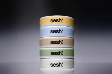 A vertical stack of five cylindrical product containers with pastel coloured bands and the Sesh+ logo set against a grey gradient backdrop.