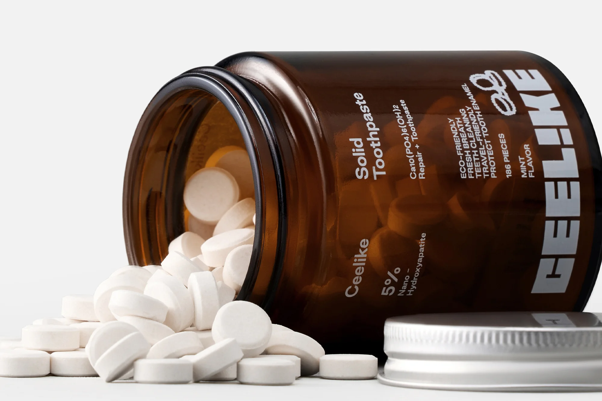 Solid toothpaste tablets spill from a brown glass jar with a silver lid, set against a clean, bright background in a product shot.