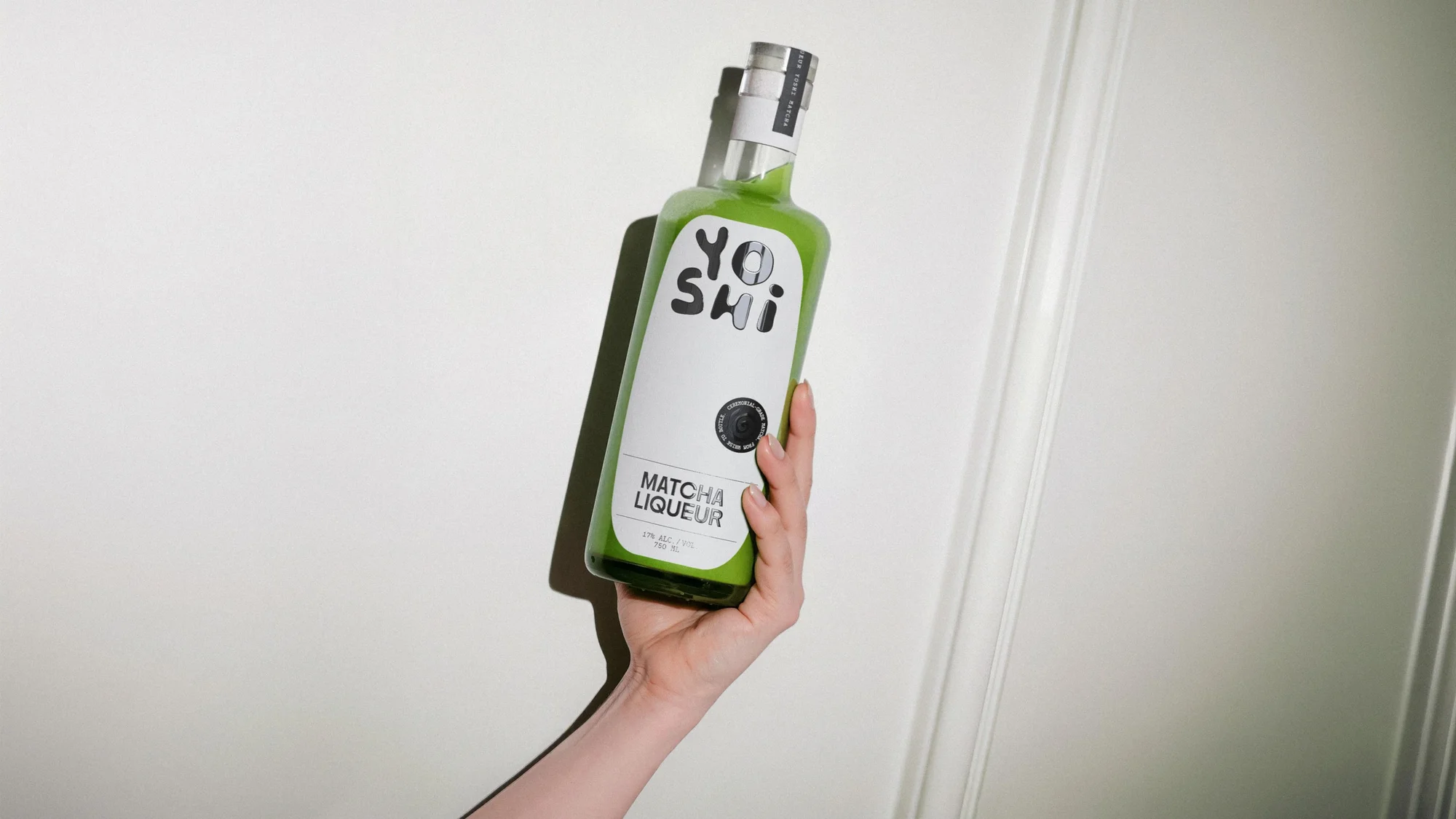 Hand holding a bottle of Yoshi Matcha Liqueur against a white wall, showcasing the product and its vibrant green color.