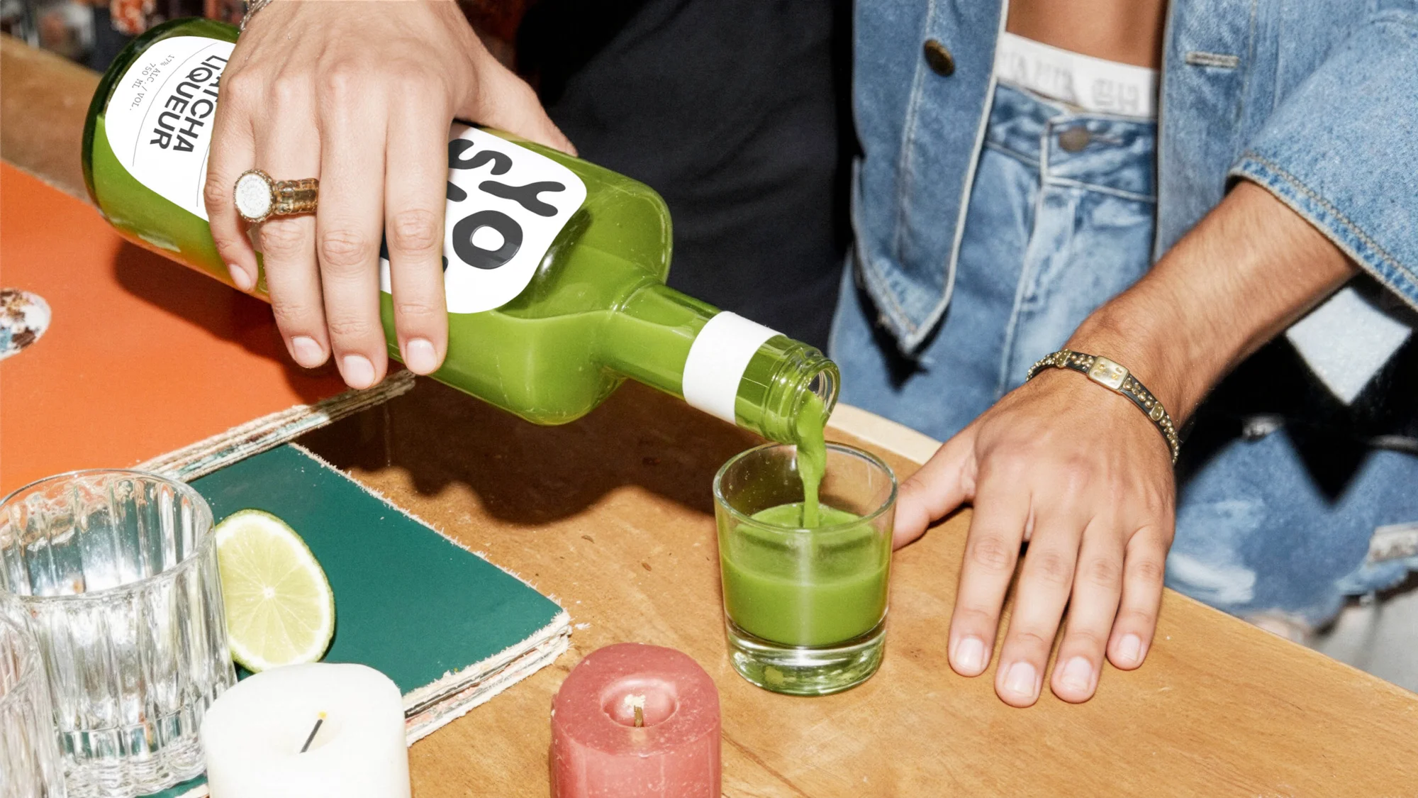 Green liquor is poured from a bottle into a rocks glass, with lime and lit candles on a wood table, showcasing a refreshing drink.