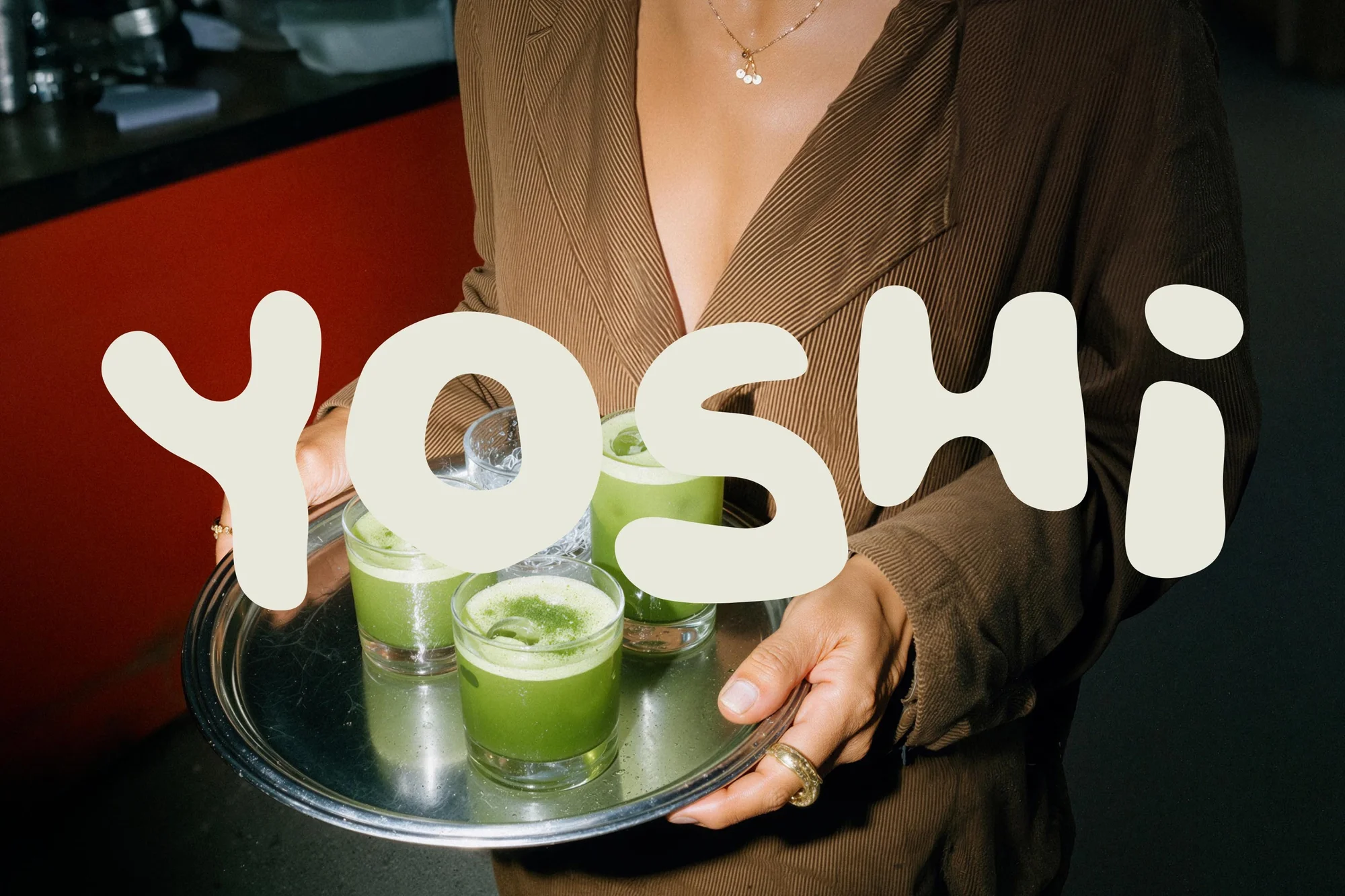 A person in a brown jacket holds a silver tray with four green drinks, under indoor lighting, the text "YOSHI" is overlaid.