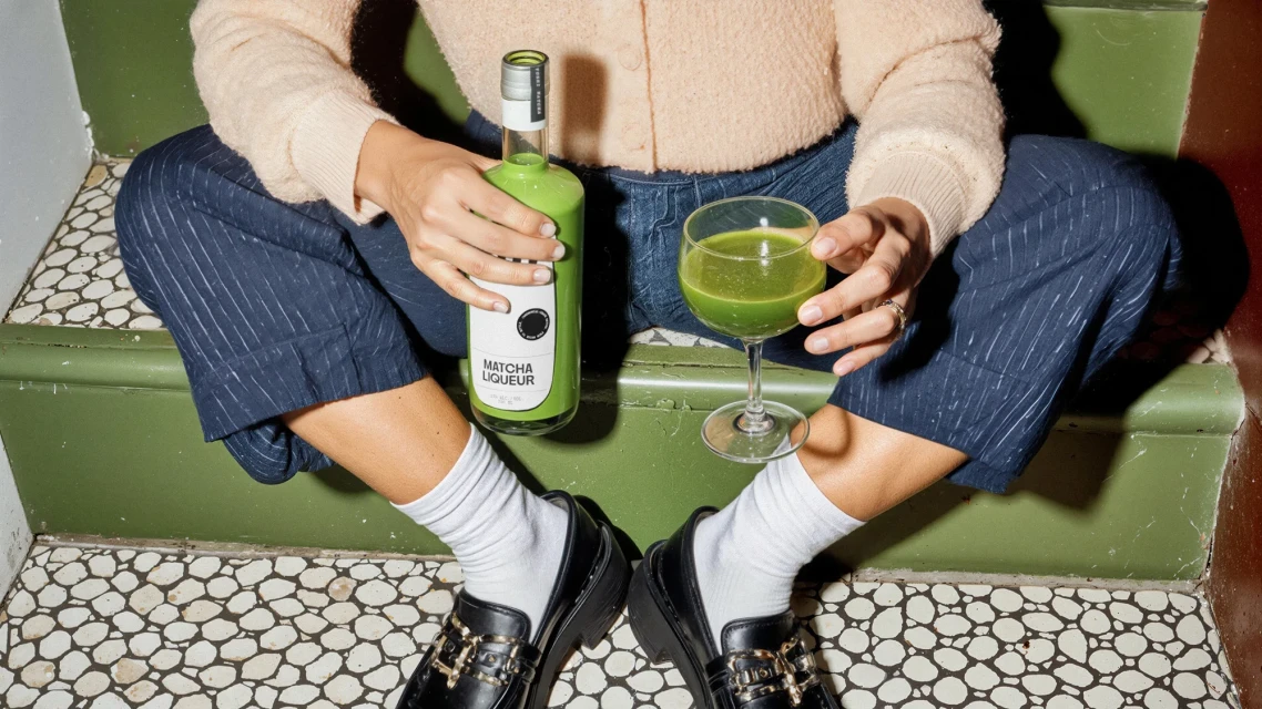 Person sits cross-legged on green steps holding a bottle of green matcha liqueur and a coupe glass filled with the same liquid.