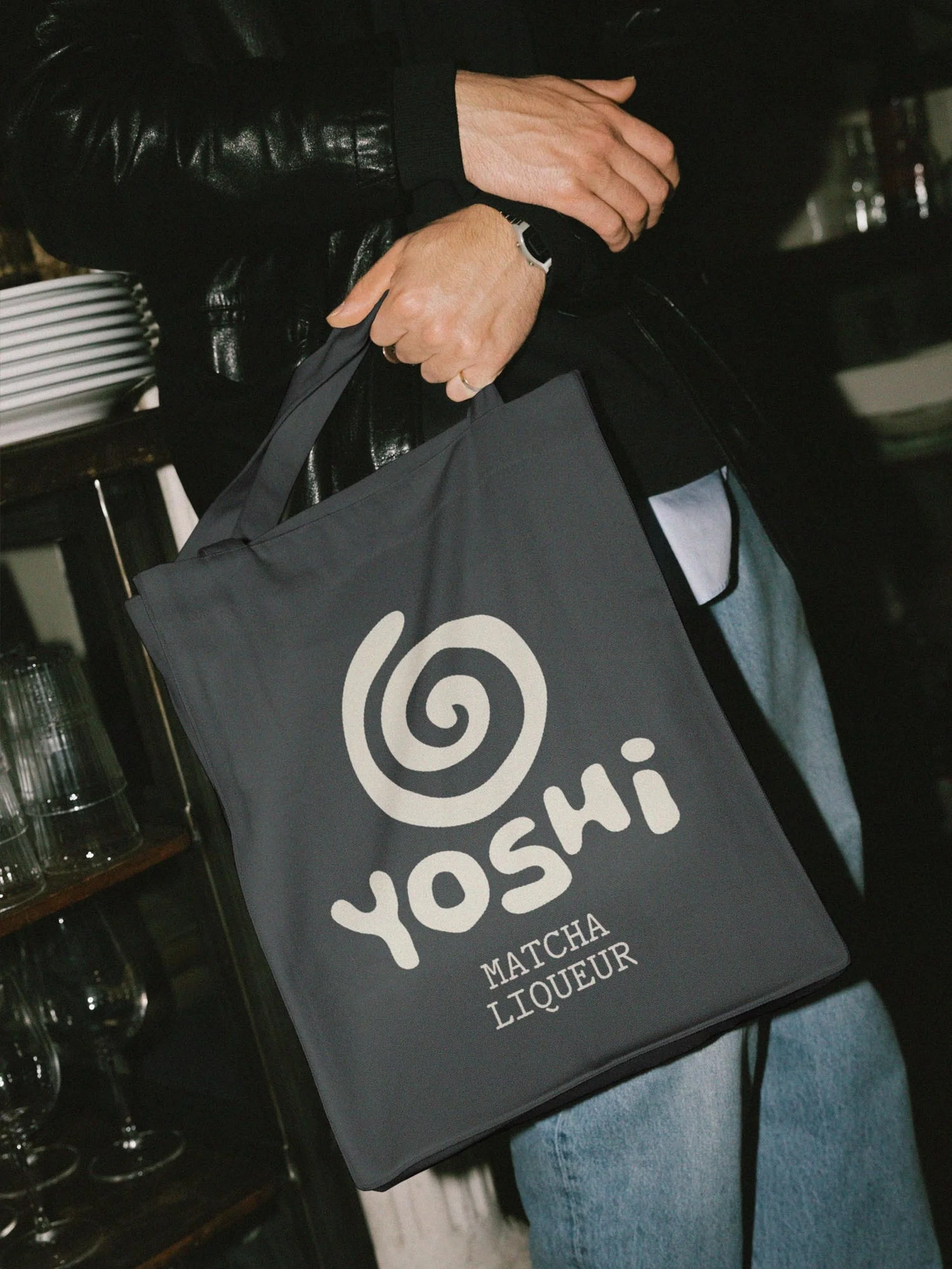 Person holding a grey tote bag with white 'YOSHI MATCHA LIQUEUR' text and logo, dressed in jeans and a black leather jacket indoors.