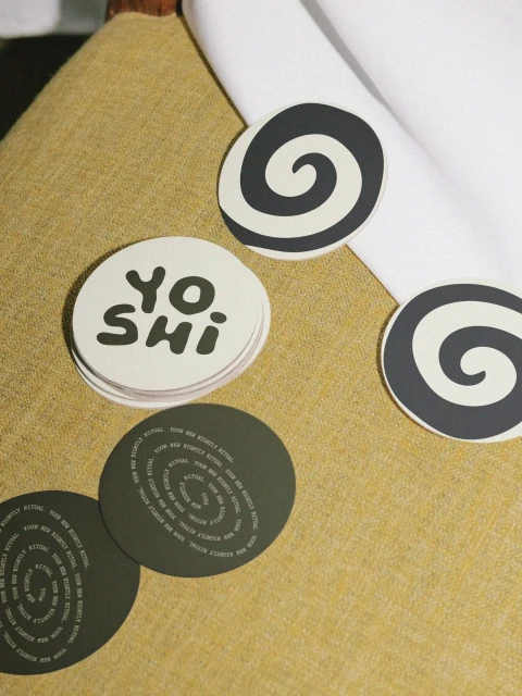 Close-up of 'Yoshi' branded and spiral-patterned drink coasters on a textured light-yellow surface, some stacked, some scattered.