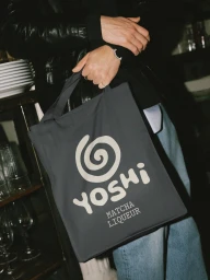 Person holding a dark grey tote bag with a white Yoshi logo and matcha liqueur text, wearing a black leather jacket.