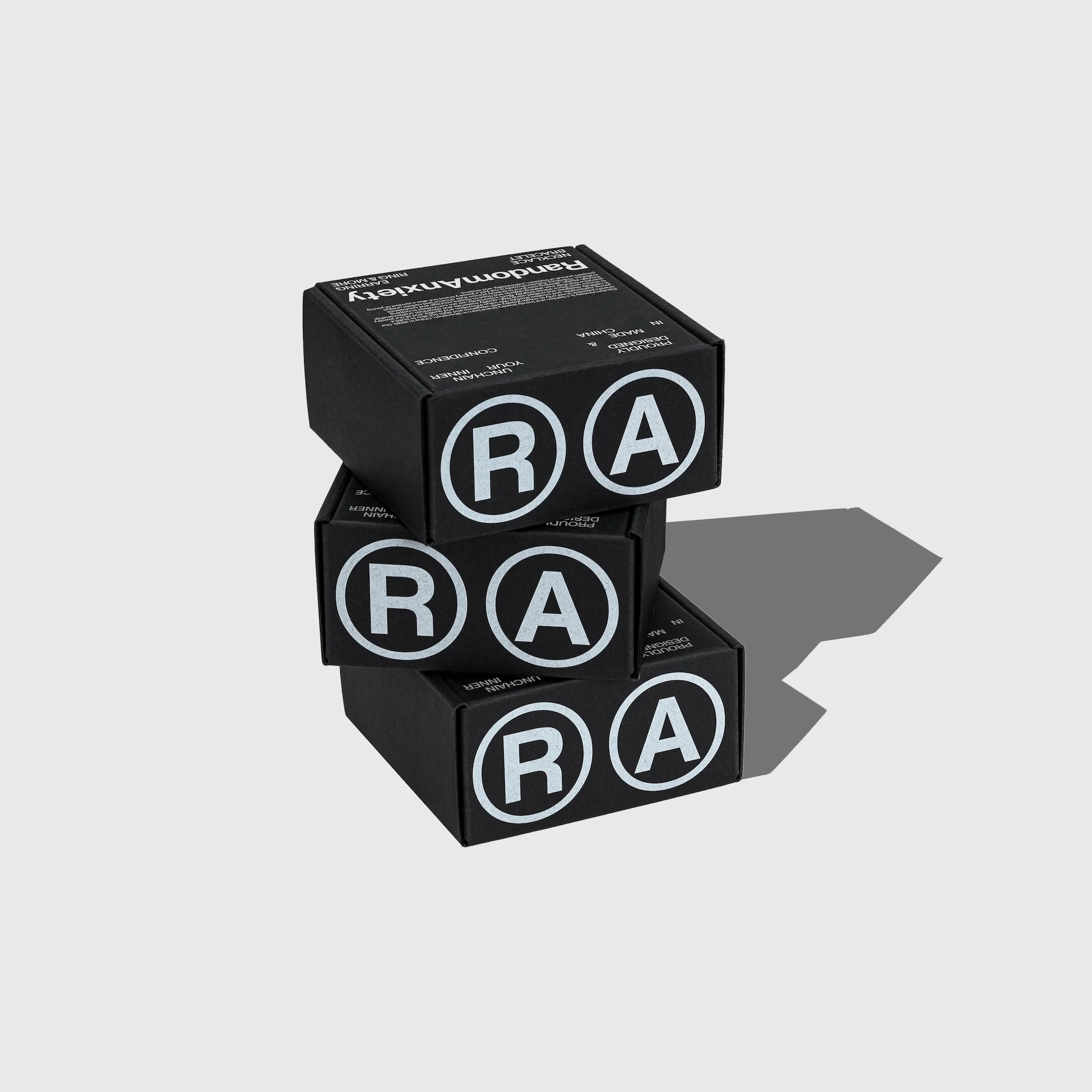 Stacked black boxes feature bold white 'RA' logo and 'Random Anxiety' text; minimalist product shot against neutral background.
