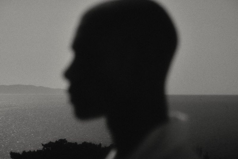 Monochrome silhouette of an adult with a blurred ocean and distant land mass in the background, evoking a sense of mystery and reflection.