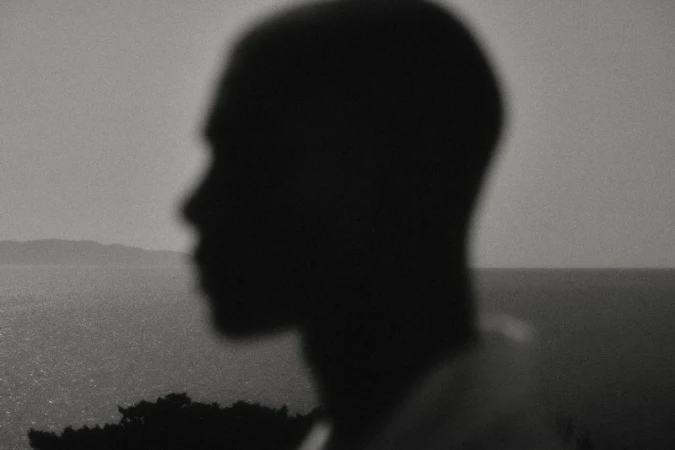 Monochrome silhouette of an adult with a blurred ocean and distant land mass in the background, evoking a sense of mystery and reflection.